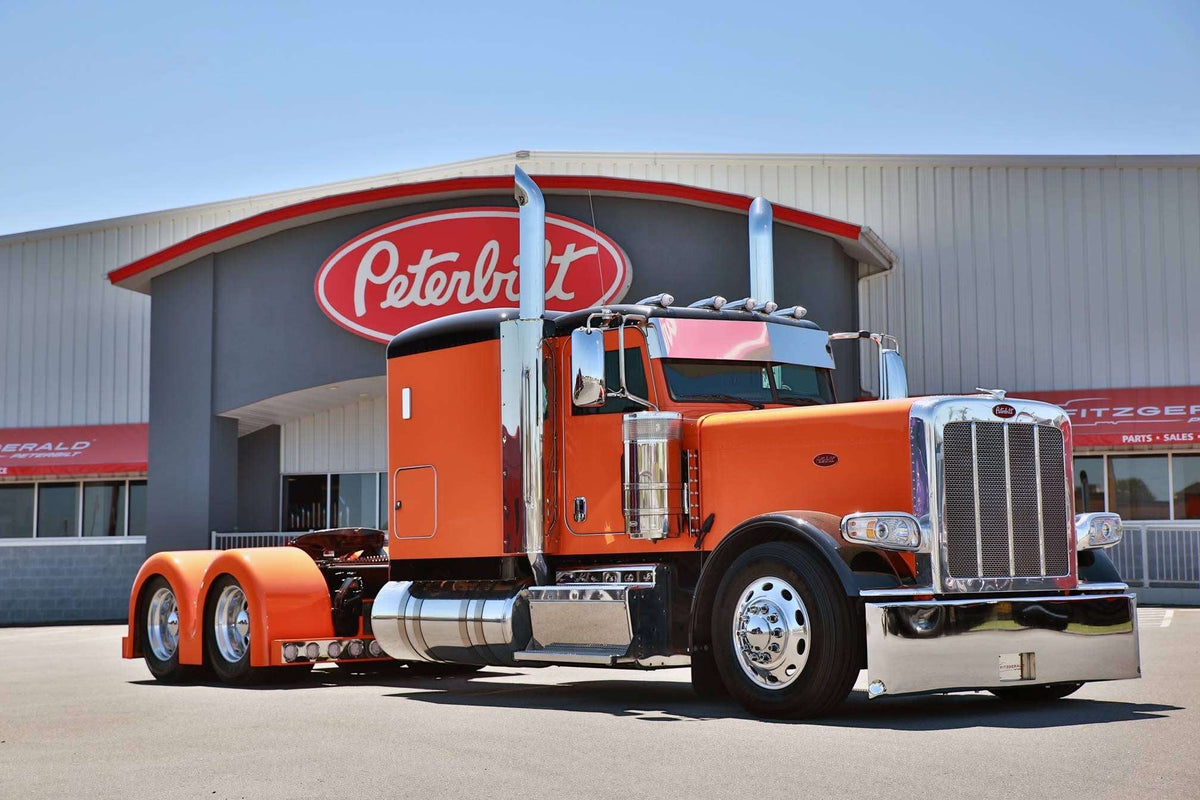 Orange Peterbilt 389 with Bad Ass double hump fiberglass rear fenders at a Peterbilt dealership